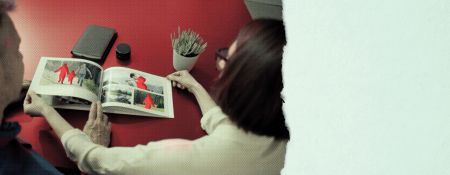 two people sit at a table looking at a school yearbook in which some of the persons are replaced by red blank outlines indicating they are no longer there